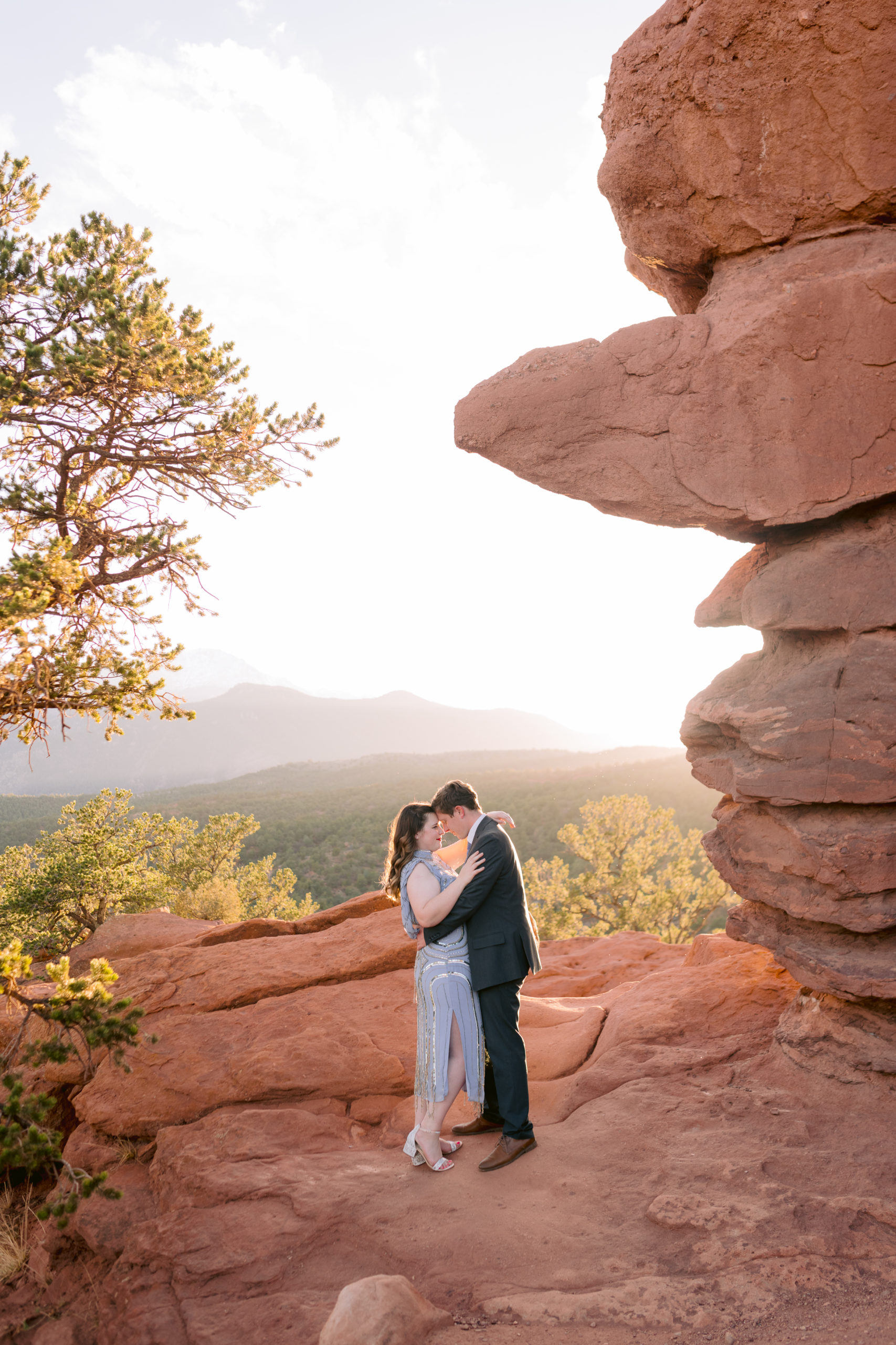 Engaged couple embraced by rock formation at Garden of the Gods overlooking Pikes Peak in the distance.